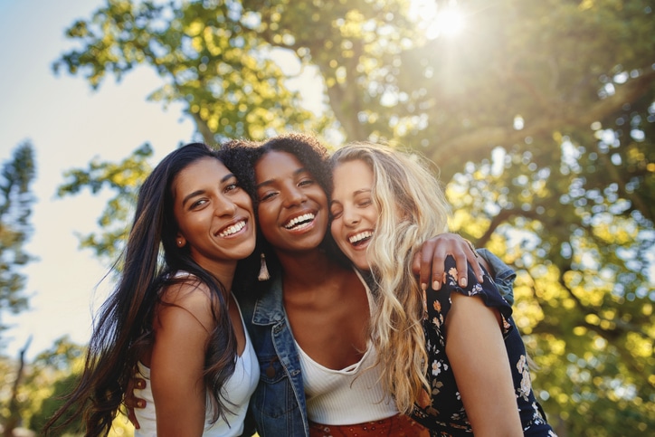 Three smiling multiethnic young women in casual wear standing together under the bright sunlight at the park having fun and laughing